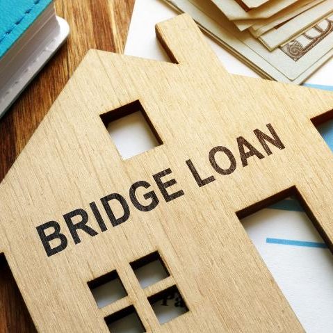 Bridging Loans Wooden house model with "BRIDGE LOAN" inscribed, surrounded by cash and a notebook.